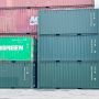 Several shipping containers stacked at a port, with visible branding and a cloudy sky in the background.