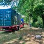 A truck with a shipping container is parked in a green area, with a person and a ladder nearby.