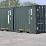 Two large shipping containers are parked in a lot, surrounded by greenery and other containers in the background.