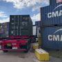 A truck with a shipping container navigates a port area filled with stacked cargo containers.